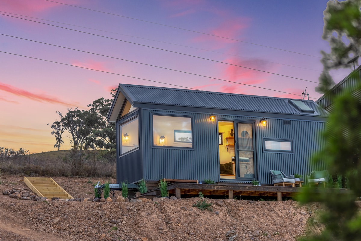 A custom-designed tiny home is showcased against a colorful sunset sky. The exterior features sleek blue metal cladding and large windows, allowing natural light inside. Steps lead up to the entrance, flanked by potted plants and outdoor seating, highlighting the inviting facade.