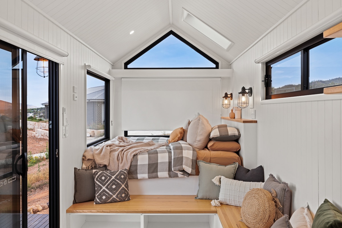 A cozy sleeping area is highlighted by a spacious bed covered with various pillows and a soft blanket. Natural light filters through large windows on both sides, enhancing the room's inviting ambiance. Simple wooden shelves provide additional decor and functionality.