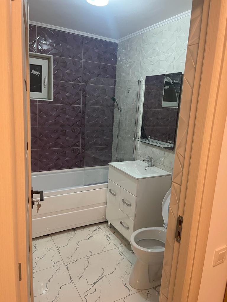 A modern bathroom is visible, featuring a shower and a bathtub combination against a backdrop of textured purple and white tiles. A mirrored cabinet is mounted above a sleek white vanity with storage drawers, complemented by a contemporary toilet. Bright ceiling lights enhance the space.