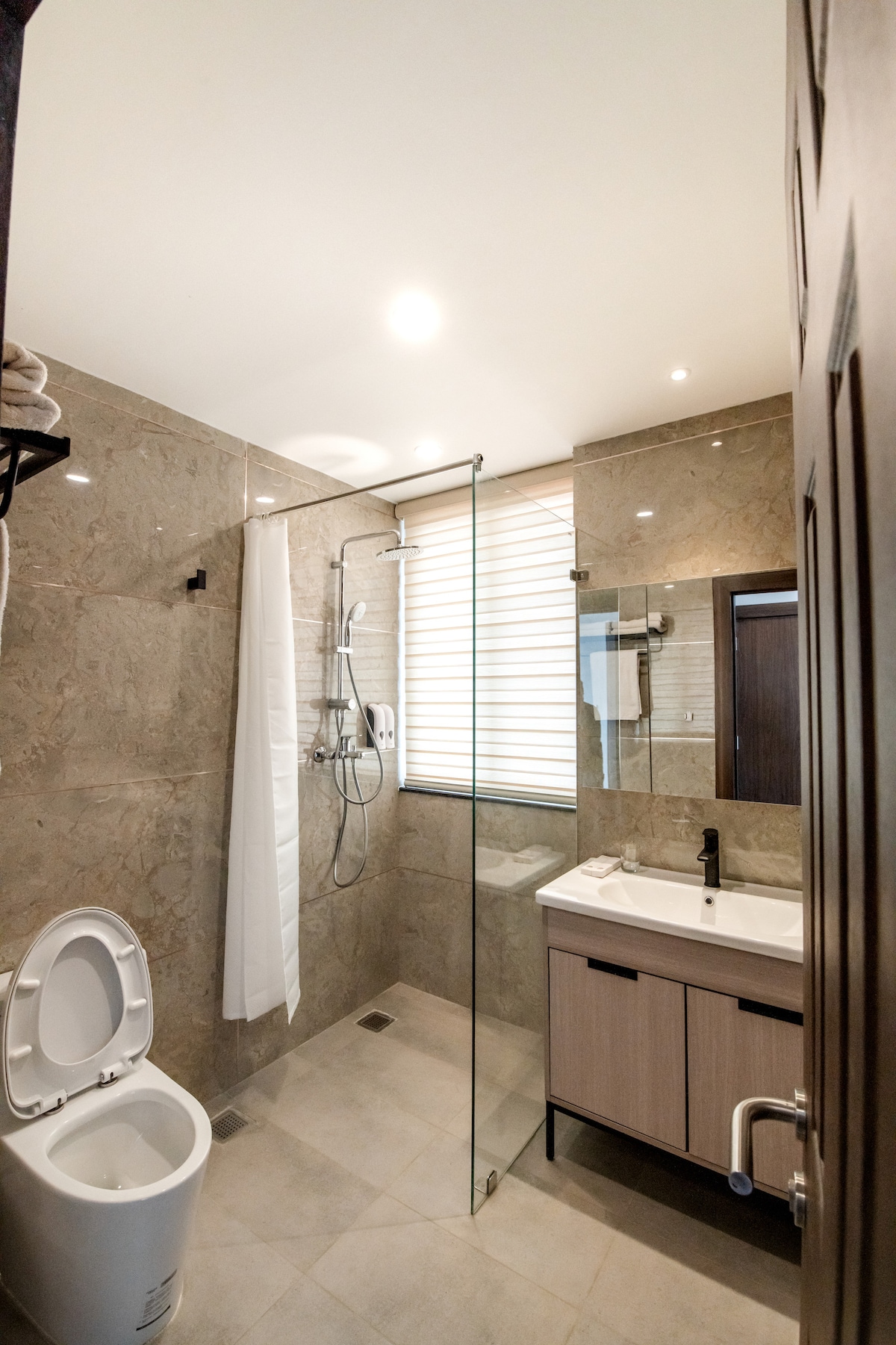 A modern bathroom is showcased, featuring a glass shower enclosure with a rainfall showerhead and a light-colored vanity with a sink. Natural light filters through partially closed window blinds, illuminating the elegantly tiled walls and floor. A white toilet is positioned neatly in the corner.