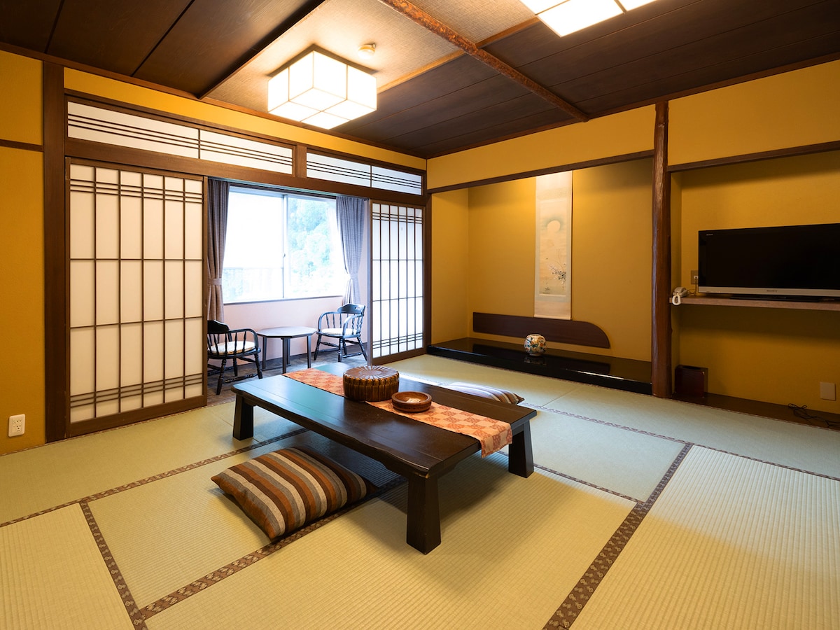 A traditional Japanese-style room features tatami flooring and sliding shoji doors. A low wooden table is set in the center, surrounded by floor cushions. Natural light filters through the window, illuminating the space. A small seating area with two chairs is visible beside the window.