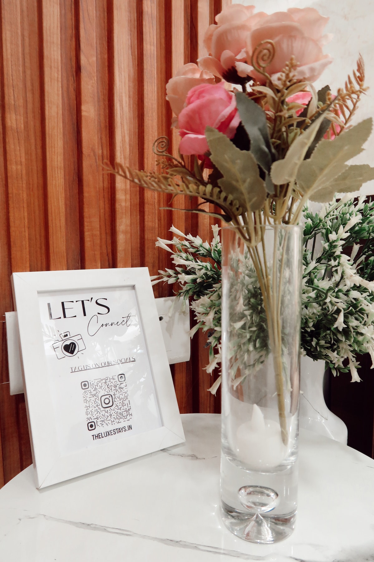 A decorative arrangement featuring a clear glass vase filled with faux flowers. The vase is placed on a white marble table beside a framed sign that invites guests to connect, featuring a QR code. Light wood paneling serves as a warm backdrop.