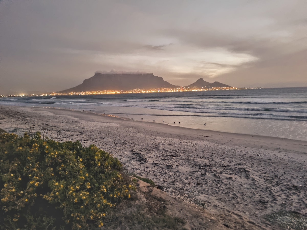 The image captures a tranquil beach scene at dusk, with the silhouette of Table Mountain and Lion's Head visible in the distance. Gentle waves lap at the shore, while faint lights from the city glow along the coastline. A patch of yellow flowers can be seen in the foreground.