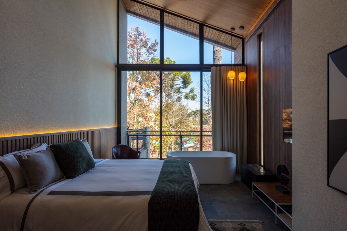 A modern suite features a spacious bed with quality bedding, complemented by large windows that provide natural light and showcase tree views. A cozy seating area includes a chair, while a freestanding bathtub is positioned near the windows, creating an inviting and serene atmosphere.