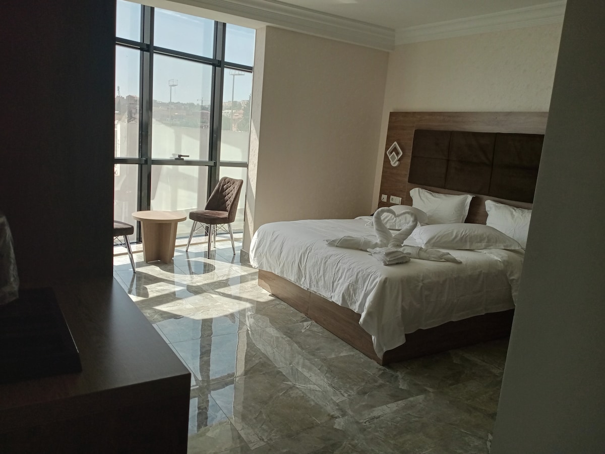 A bright and airy sleeping area features large windows allowing natural light to fill the space. A spacious bed is situated against a natural wood headboard, accompanied by two elegantly arranged chairs and a round table. Marble flooring complements the modern aesthetic.