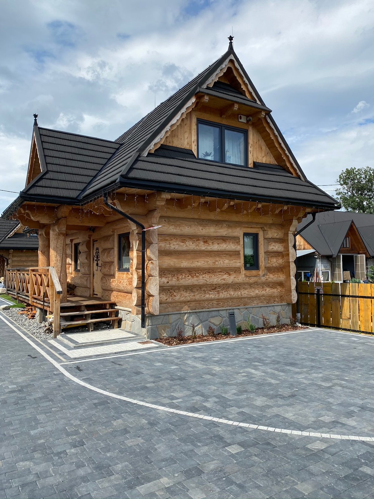 A charming wooden house features a traditional log cabin design with a dark roof and large windows. Nestled in a scenic area, the inviting front porch showcases wooden railings and steps leading to the entrance, surrounded by a landscaped yard and paved driveway.