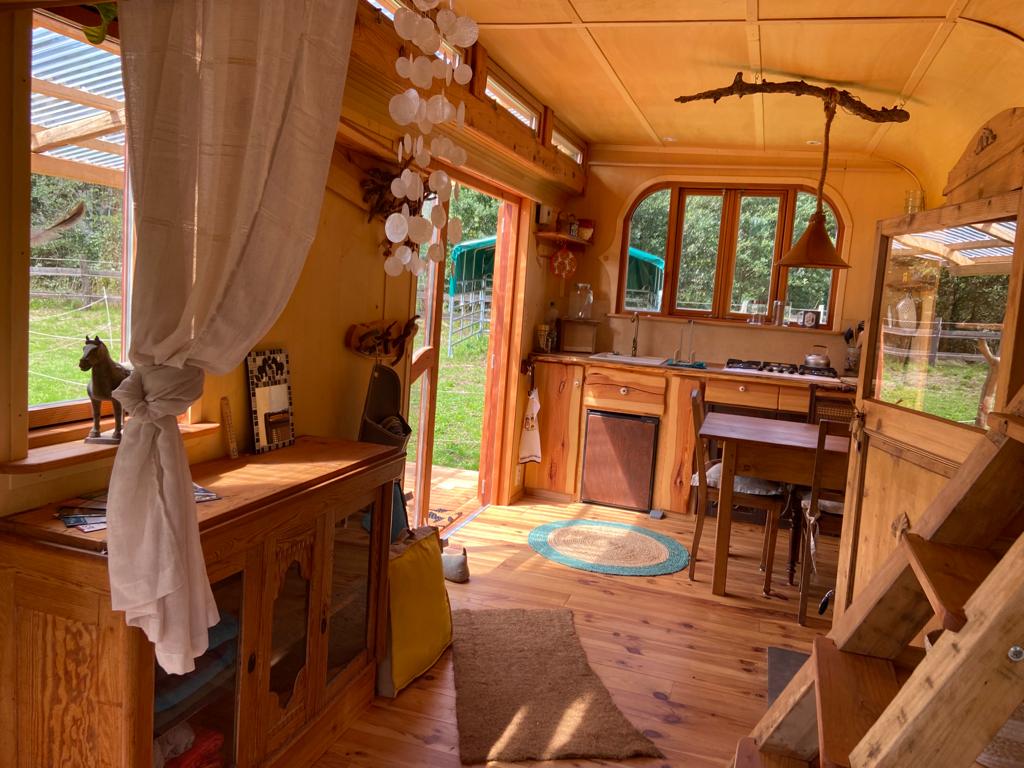 The interior of the Tinyhouse features warm wooden cabinetry and a cozy seating area. Natural light floods in through large windows. A compact kitchen with a gas stove is visible, alongside a dining table. Decorative elements, including hanging lanterns, add charm to the space.