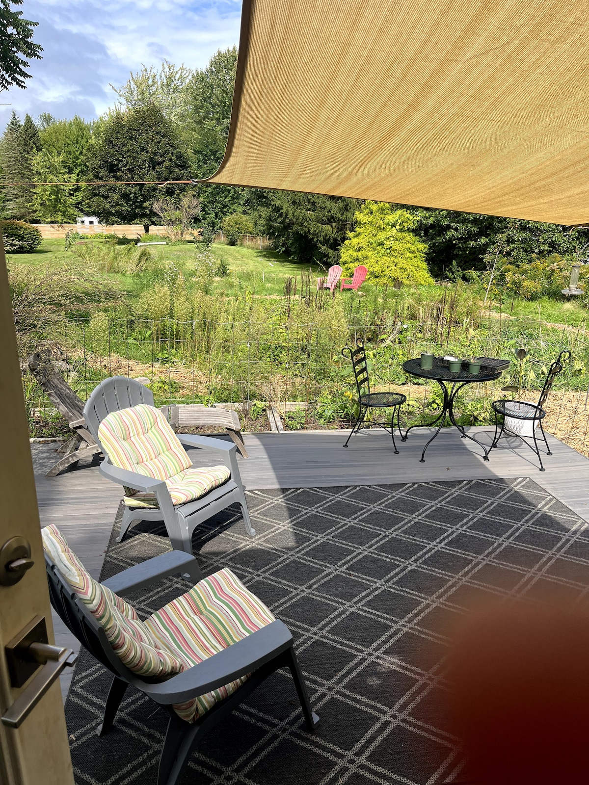 A spacious deck is visible, featuring comfortable seating with striped cushions. A small round table and chairs provide a cozy gathering space. In the background, a lush yard filled with wildflowers and greenery is surrounded by trees, creating a tranquil outdoor setting.