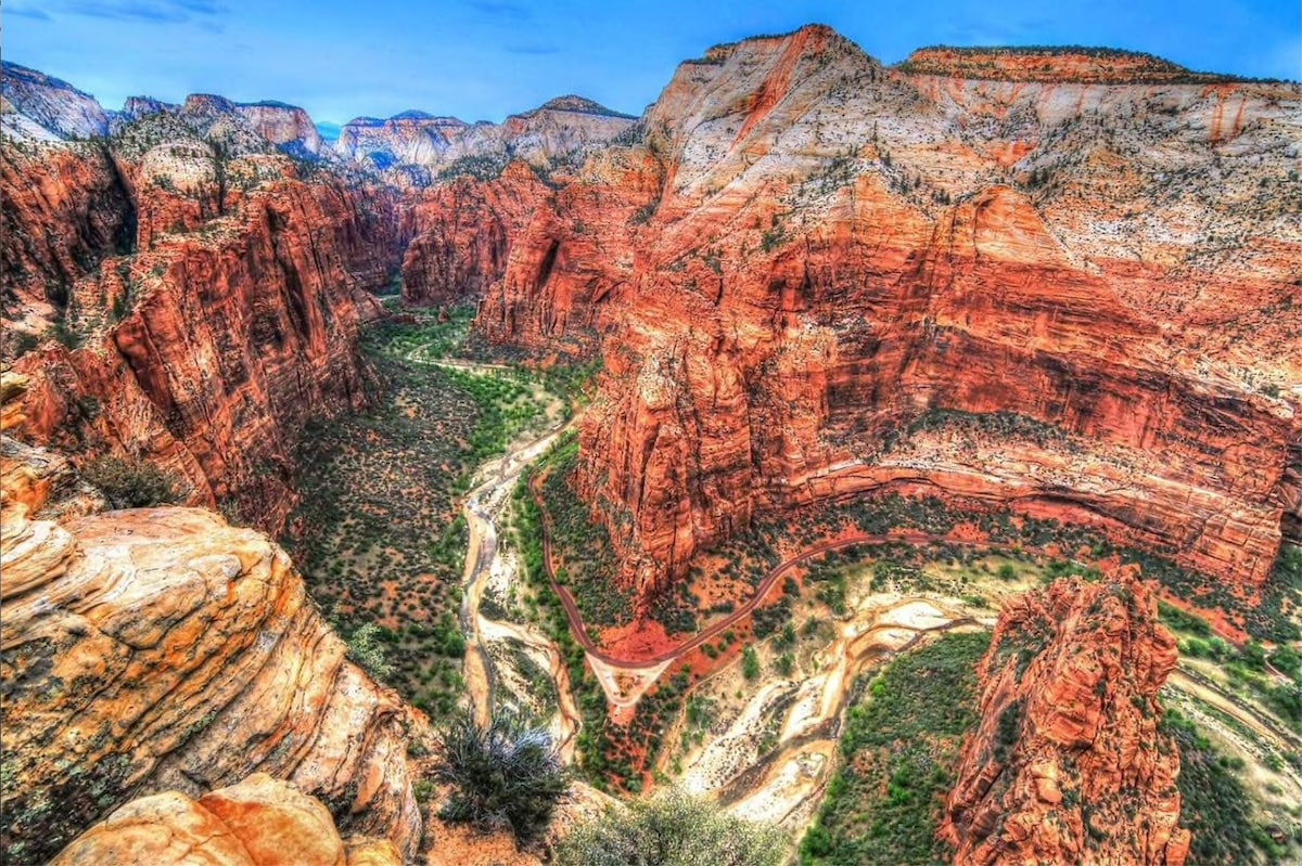 A breathtaking view of the vast Zion Canyon showcases the striking red and orange rock formations that characterize the area. Lush greenery lines the canyon floor and the Virgin River meanders through the rugged landscape, framed by towering cliffs and scenic vistas under a clear blue sky.