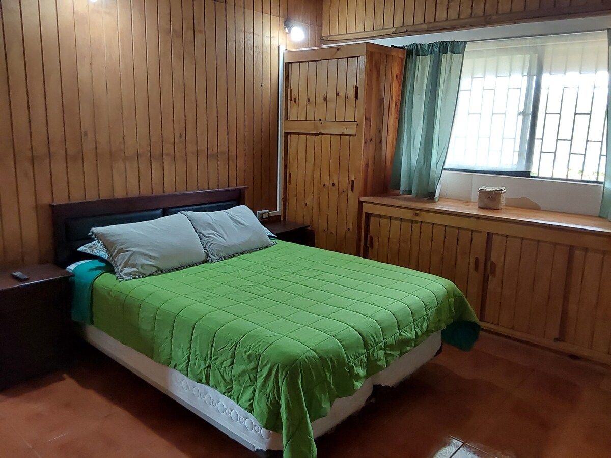 A spacious sleeping area is shown, featuring a green quilt covering a large bed with two pillows. Wooden walls and furnishings create a warm ambiance. Natural light filters through the window, adorned with light curtains, illuminating the room's inviting features.