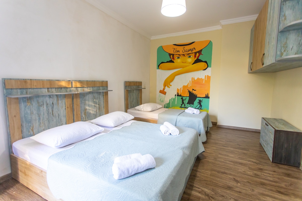 A spacious sleeping area features two beds covered with light blue bedspreads. Decorative pillows are placed at the head of each bed. A vibrant mural is displayed on one wall, adding a pop of color to the neutral decor. Light wood furniture complements the room's design.
