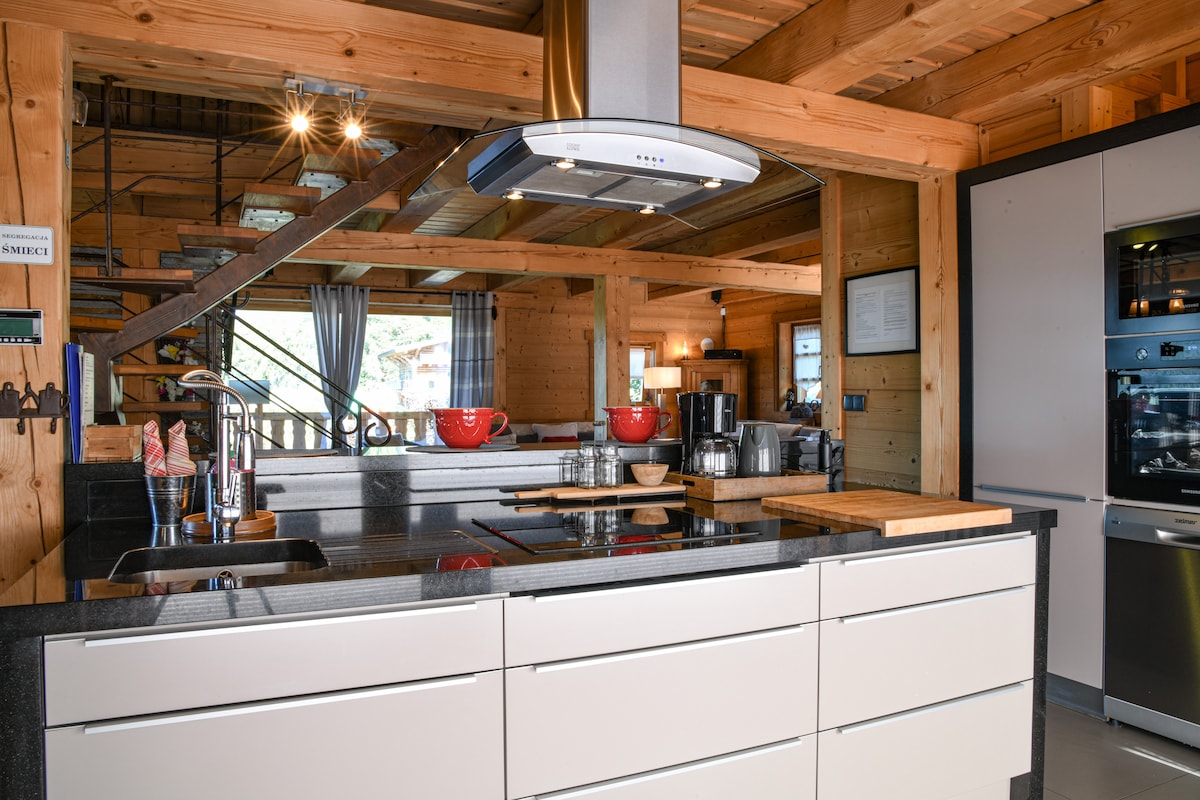 The kitchen features a modern design with a central island, equipped with a sleek countertop and a range hood above. Stainless steel appliances are present, complemented by wooden accents throughout the space. Natural light enters from the large windows, enhancing the functional layout.