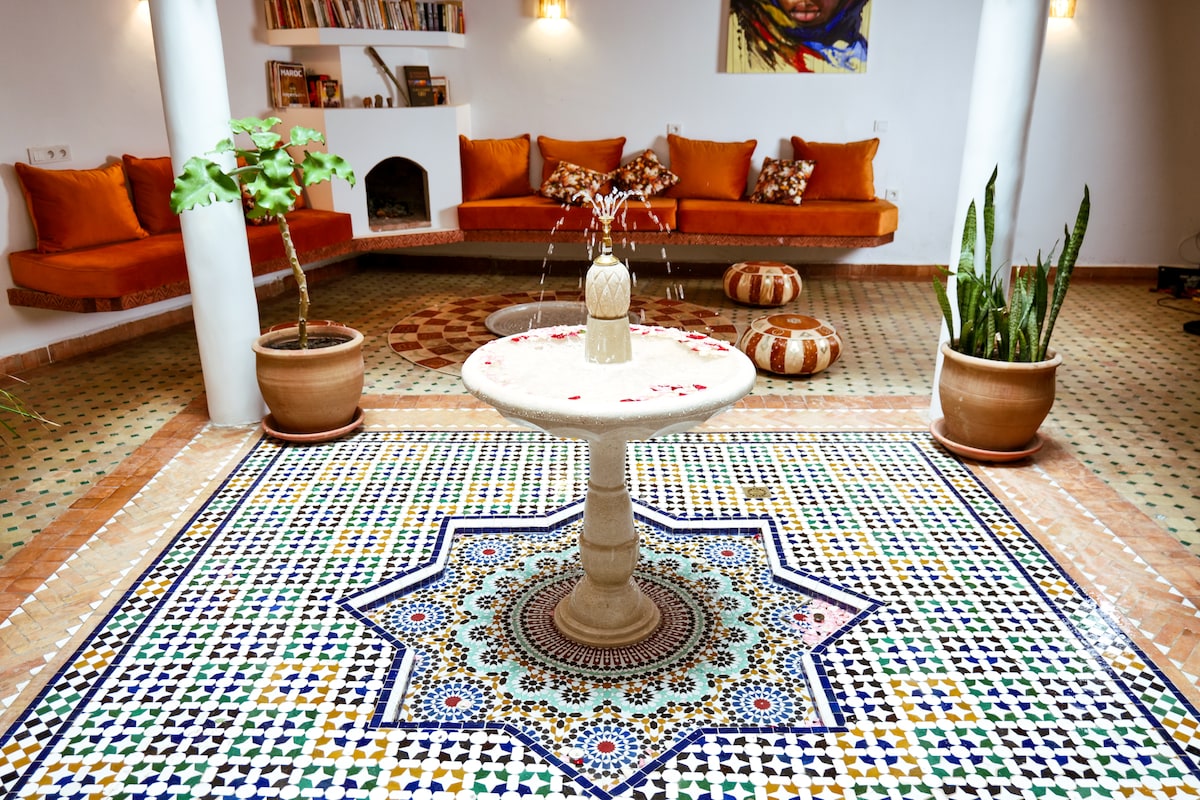A central fountain is surrounded by intricately patterned tiles, showcasing a vibrant mosaic design. The seating area features orange cushions against a backdrop of neutral walls. Potted plants are positioned nearby, adding a touch of greenery to the inviting space.