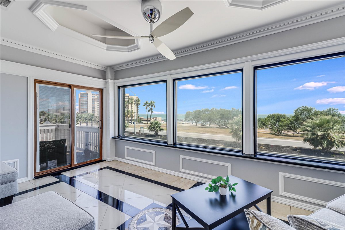 A sunroom with large windows providing expansive views of the outdoor scenery, including palm trees and open space. The room features a sleek ceiling fan and modern flooring, complemented by a simple green plant on a small table.