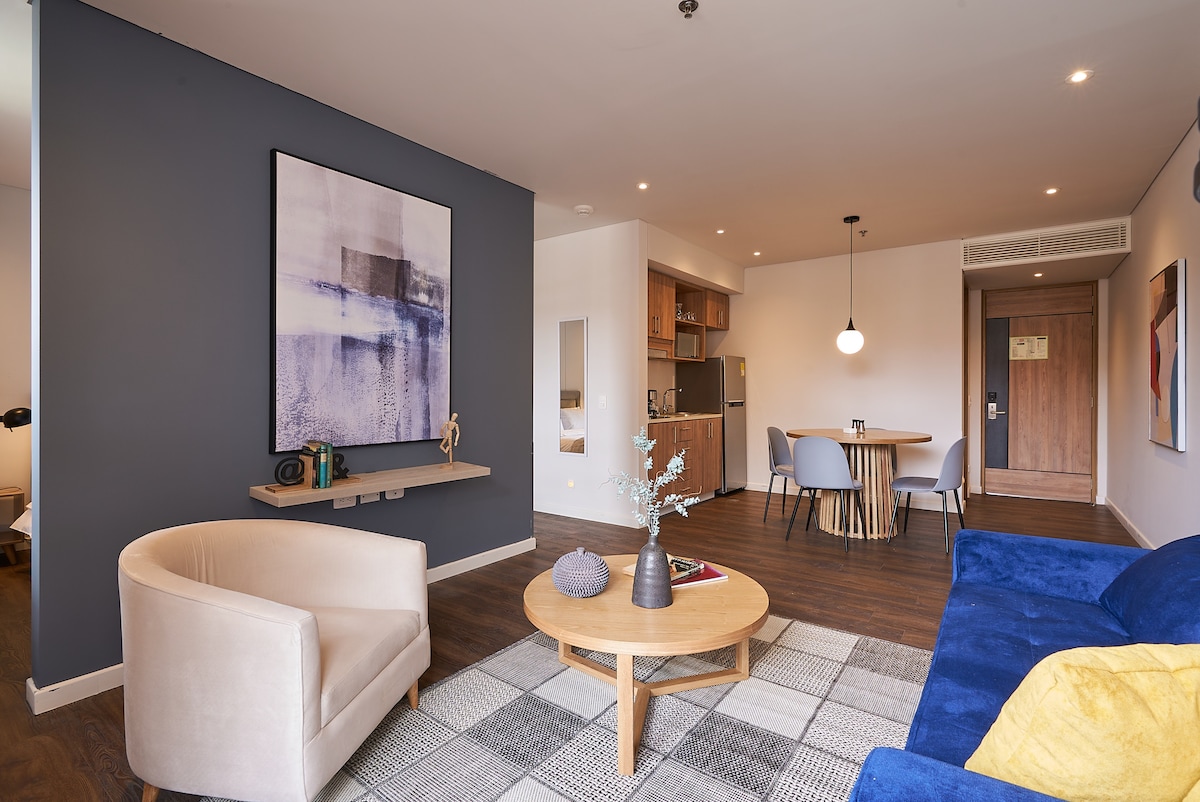 A modern living area featuring a cozy blue sofa and beige armchair is centered around a round wooden coffee table. A patterned rug adds warmth to the space, while a dining area with a round table is illuminated by a pendant light. Neutral artwork and decorative elements enhance the decor.