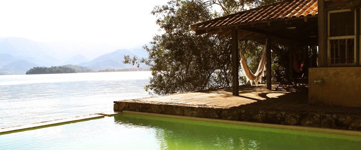 A tranquil outdoor area features a sparkling pool with a gentle green hue, bordered by wooden decking. A shaded seating area with a hammock invites relaxation, while the serene waters and distant mountains create a peaceful backdrop.