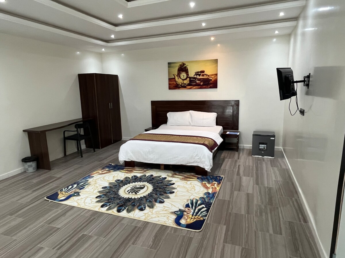 A spacious room features a large bed with crisp white linens, accompanied by a decorative rug with floral patterns. A wooden wardrobe and a small desk with a chair are situated nearby. The wall holds a flat-screen TV and a framed artwork, enhancing the overall ambiance.