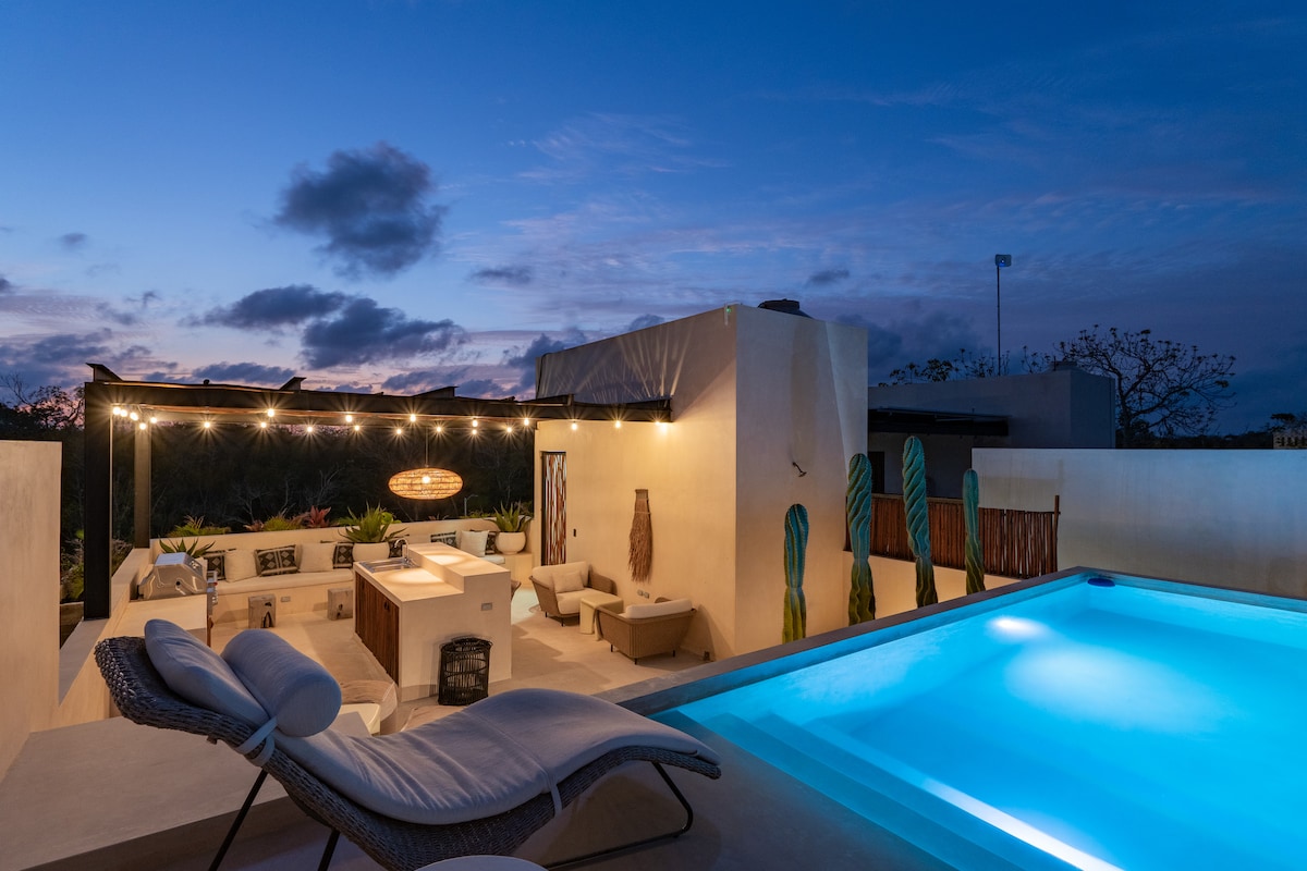 A serene outdoor space features a private pool illuminated in soft blue light. Comfortable seating areas are arranged near a barbecue station, surrounded by lush greenery. String lights add a warm ambiance to the area, creating a welcoming atmosphere for relaxation.