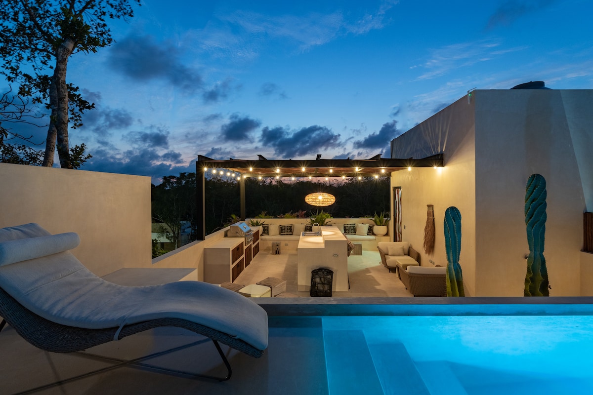 A private patio area overlooks a tranquil scene, featuring a pool and lounge chair. String lights create a warm ambiance, while an outdoor kitchen and seating area are visible. Lush greenery complements the surroundings, enhancing the connection to nature.