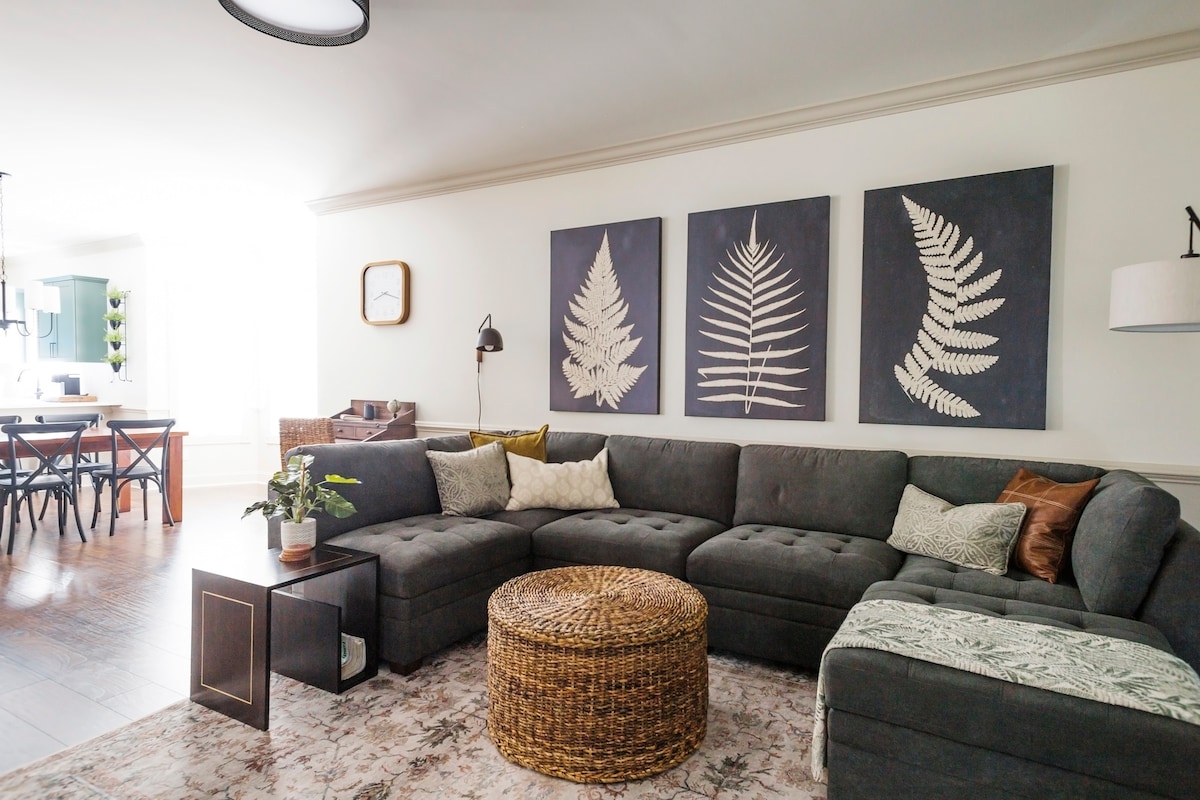 The living area features a large sectional couch with accent pillows and a round woven coffee table. Three framed botanical prints adorn the wall, while light enters through a nearby window. A floor lamp and decorative items enhance the inviting ambiance.