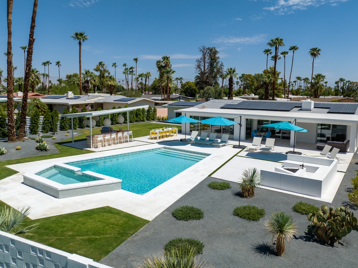 The outdoor area features a spacious saltwater pool surrounded by lounge chairs and colorful umbrellas. A firepit seating area and an outdoor dining space are visible, framed by palm trees and landscaped greenery. The home showcases a modern design with expansive windows.
