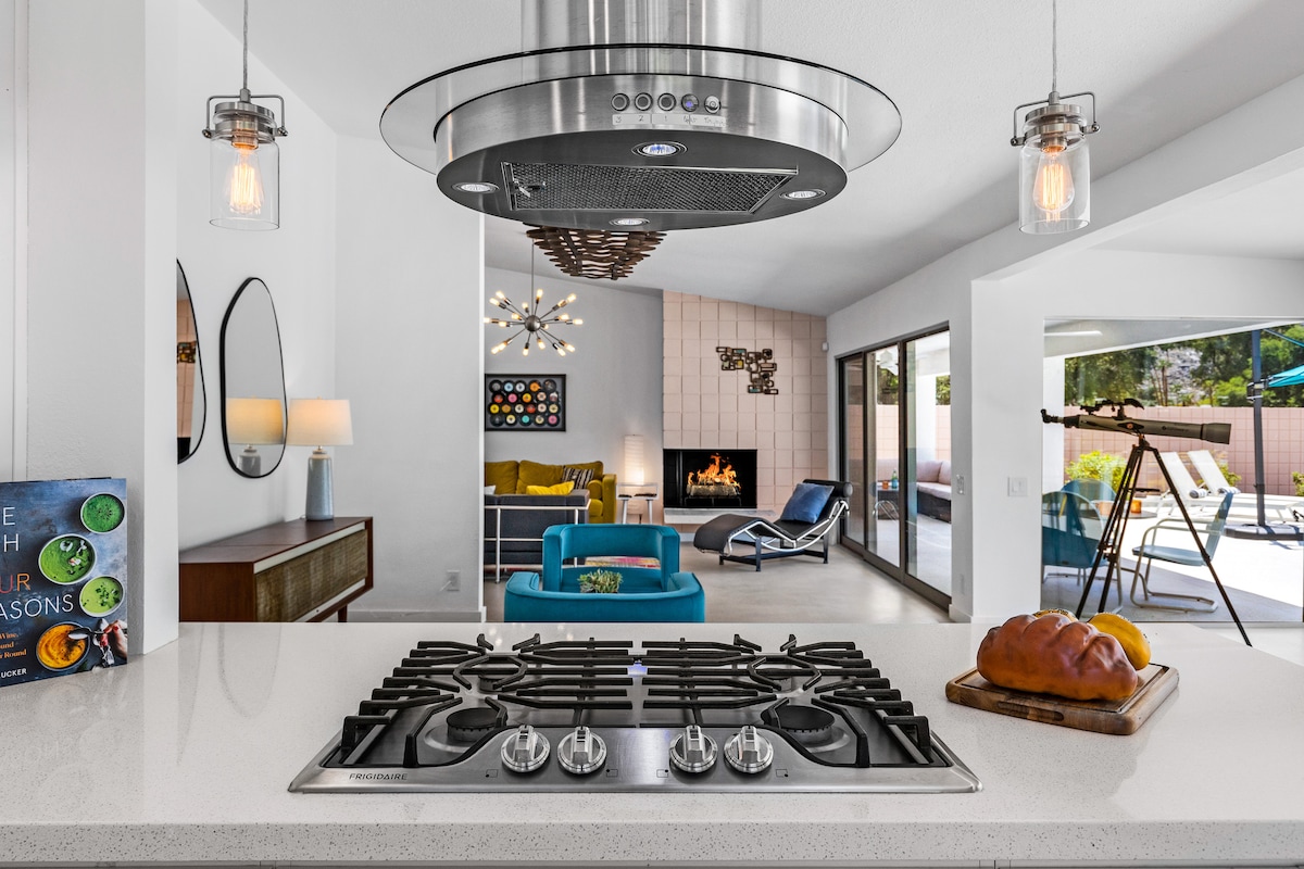 A modern kitchen with a sleek gas stovetop is visually connected to a spacious living area. The space features stylish lighting fixtures and a fireplace, while large glass doors provide a view of the outdoor area with seating and a pool.