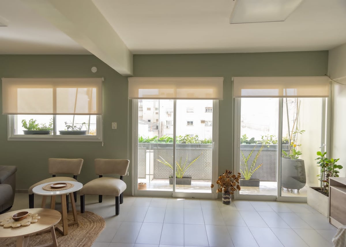 A bright living space is presented with two large glass doors leading to a balcony. Neutral-colored walls complement the light tile floor. Two chairs are positioned beside a circular table, while potted plants decorate the balcony, adding a touch of greenery.