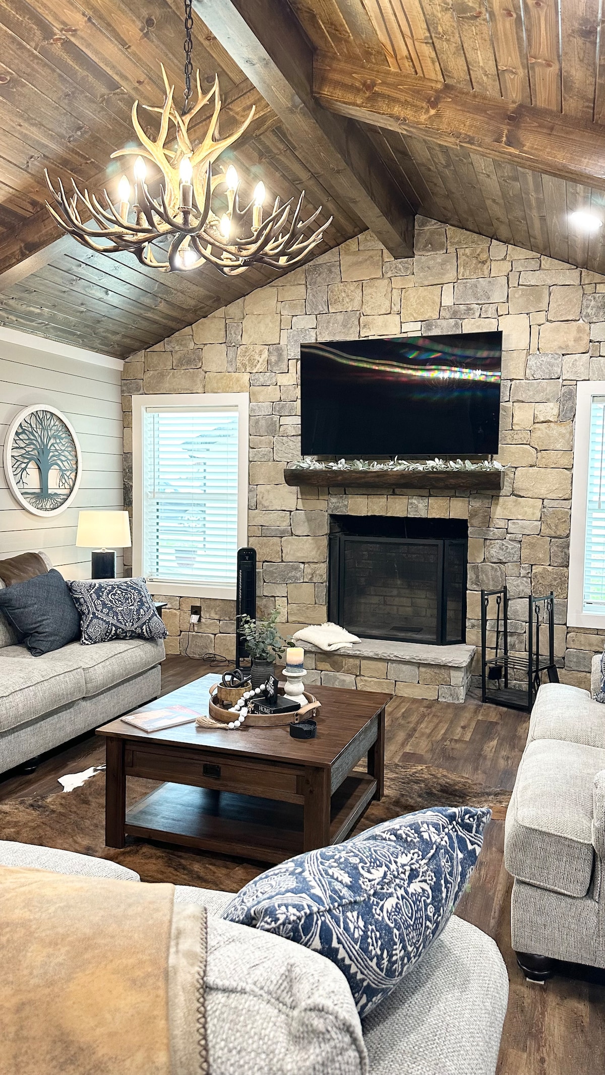 The living room features a stone fireplace surrounded by wood accents and a large smart TV mounted above. Soft seating includes a cozy gray sofa and an armchair, accompanied by a wooden coffee table. A rustic chandelier hangs from the vaulted ceiling, adding warmth to the space.
