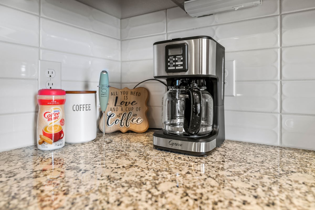 The kitchen countertop features a coffee maker alongside a container labeled 'Coffee.' A decorative wooden sign displaying the phrase 'All you need is Love and a cup of Coffee' adds a personal touch. The sparkling granite surface enhances the modern vibe of the space.