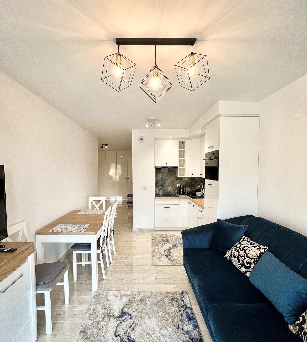 A modern living space is depicted, featuring a dark blue sofa paired with a patterned cushion. A dining table with white chairs occupies one side, while attractive light fixtures hang above. The kitchen area is visible in the background, offering a clean, functional design.