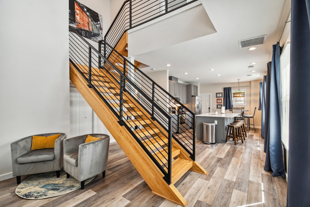 A modern interior showcases a wooden staircase with a sleek metal railing, leading to the upper level. The open living space features a cozy seating area with two chairs and a round rug. In the background, a kitchen bar with stools invites social gatherings.