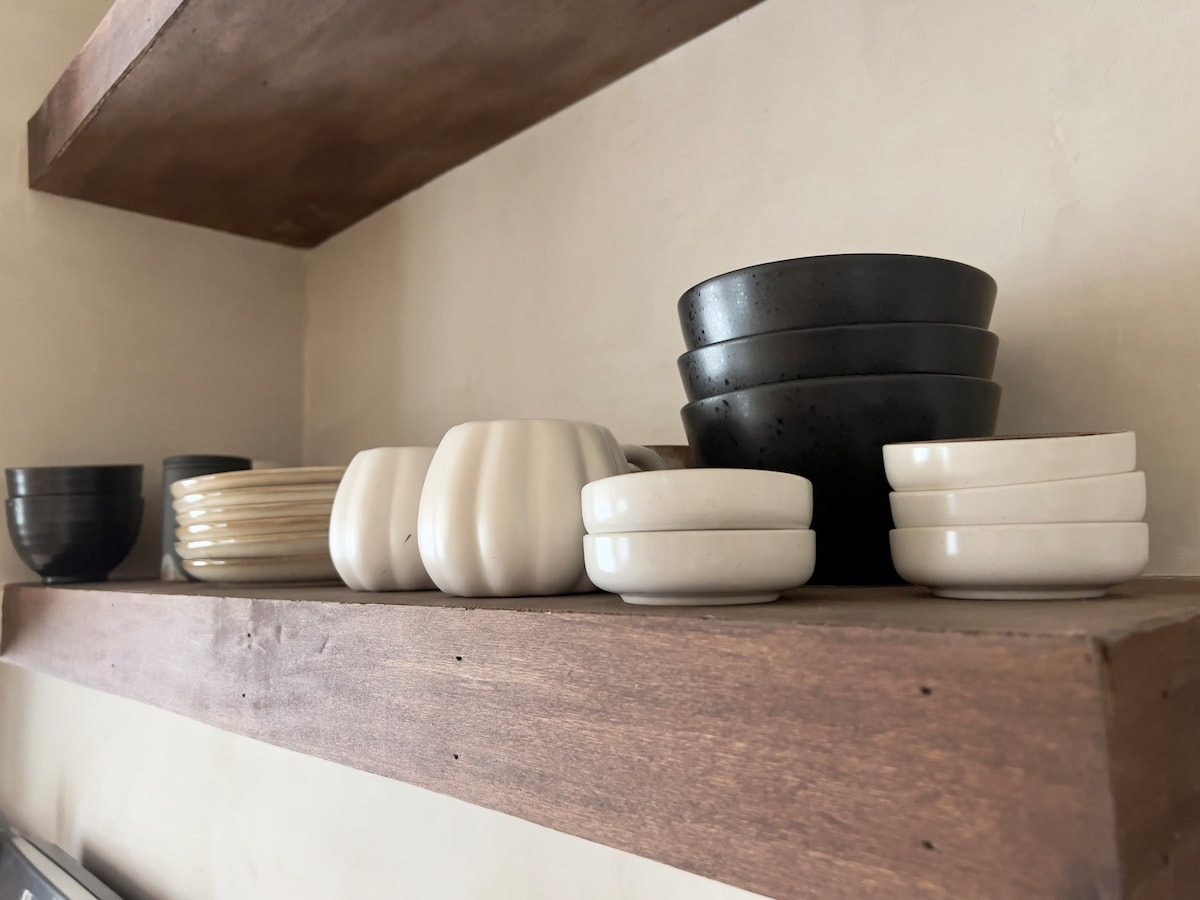 A wooden shelf displays an assortment of bowls and dishes in various shapes and colors. Black bowls sit at the back, with white and cream-colored pieces arranged in front. The simple design showcases a blend of textures and materials.