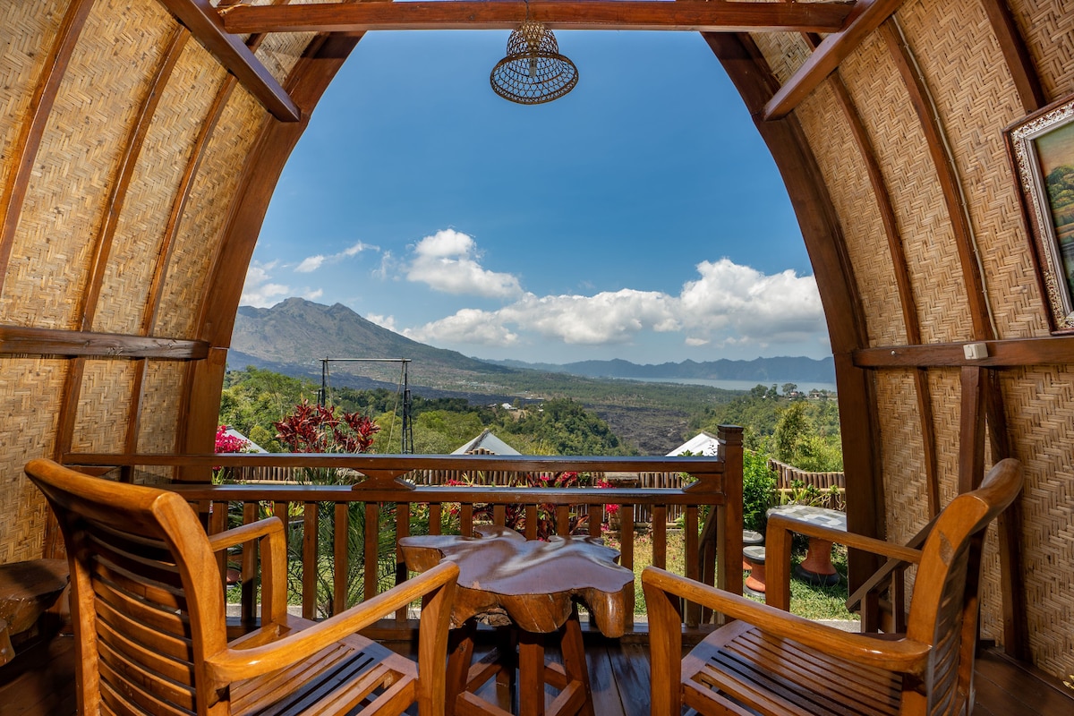 A spacious balcony features two wooden chairs and a unique wooden table, offering unobstructed views of the lush landscape and Mount Batur in the distance. Natural light fills the area, enhancing the warm tones of the bamboo walls and ceiling.