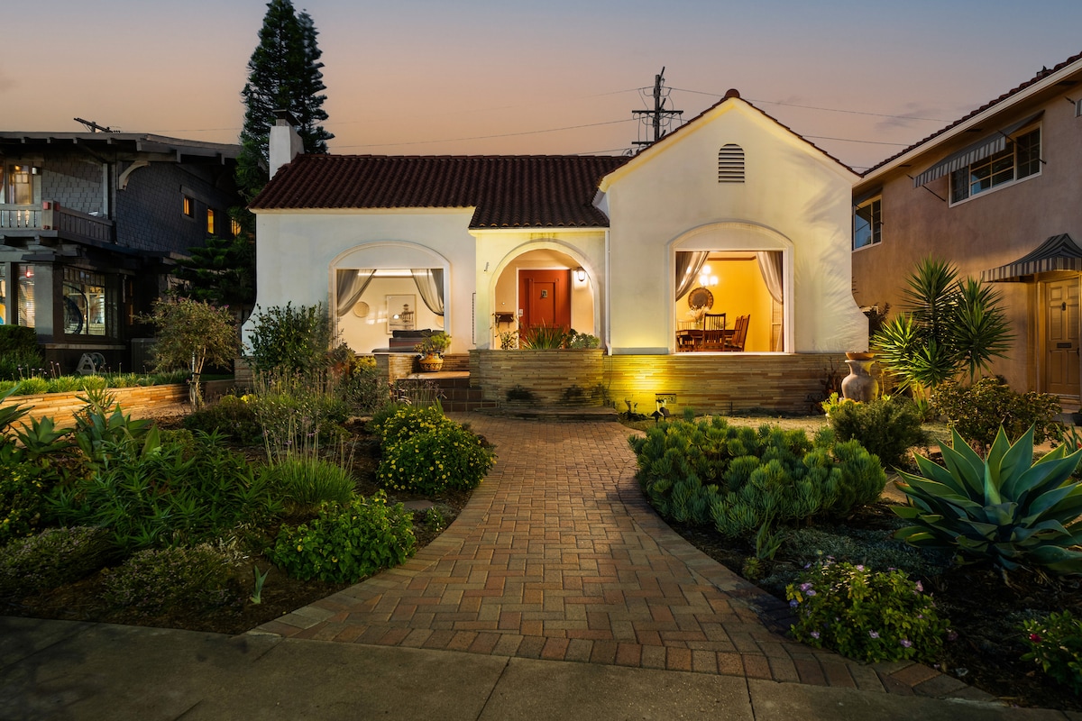 The exterior of a charming Spanish Mission Revival home is presented, featuring a welcoming pathway leading to a well-lit entrance. Lush landscaping surrounds the property, with various plants and trees framing the structure, which boasts arched windows and a warm glow from inside.
