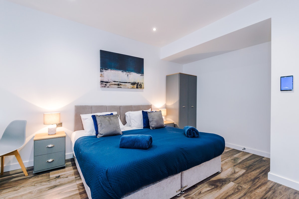 A spacious bedroom is presented with a large bed dressed in blue linens. Nightstands with lamps flank the bed, and an abstract artwork adds a contemporary touch to the decor. A wardrobe stands nearby, complementing the light wood flooring and bright ambience.
