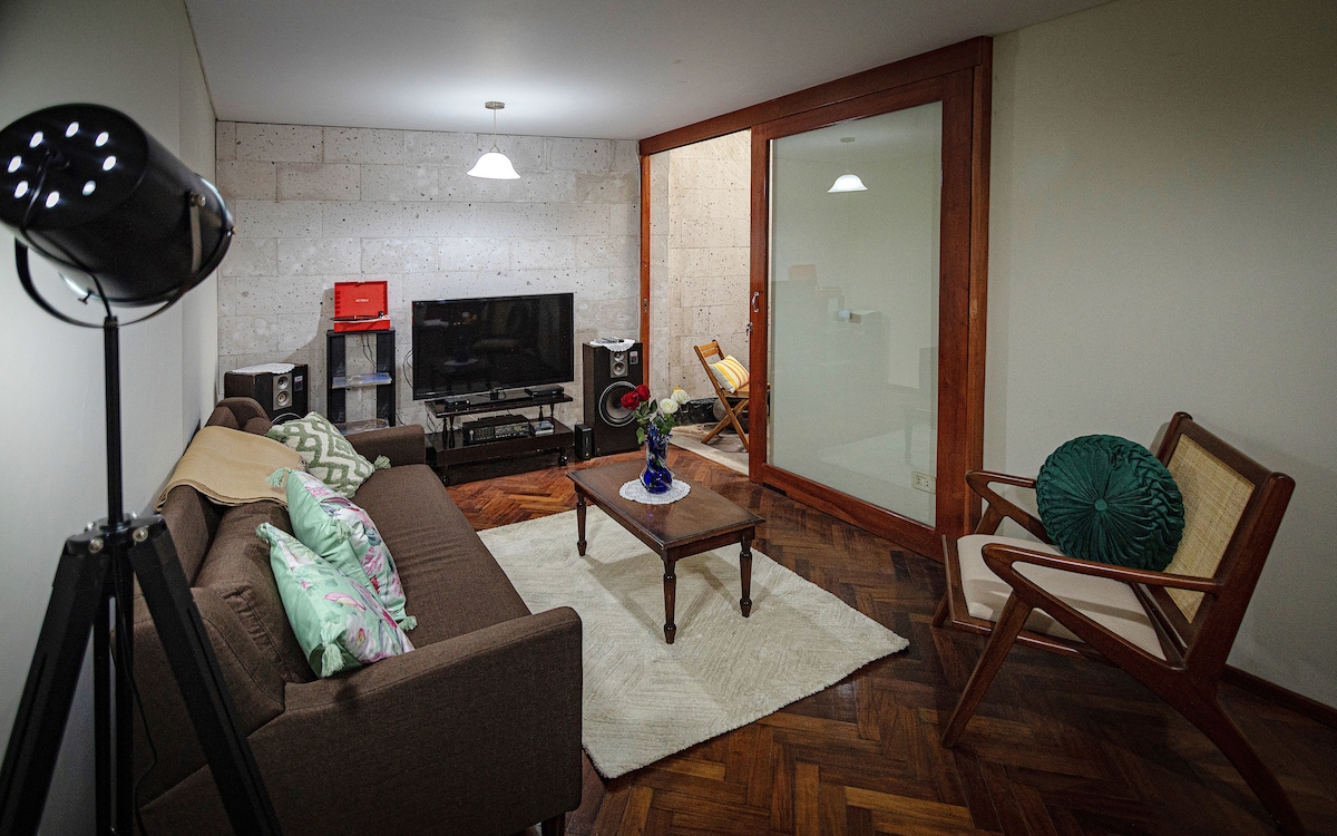 A modern living area is highlighted by a dark sofa adorned with patterned cushions. A coffee table sits on a soft rug, while a television is positioned on a wooden stand. Glass doors provide access to a separate space, enhancing the room's natural light.