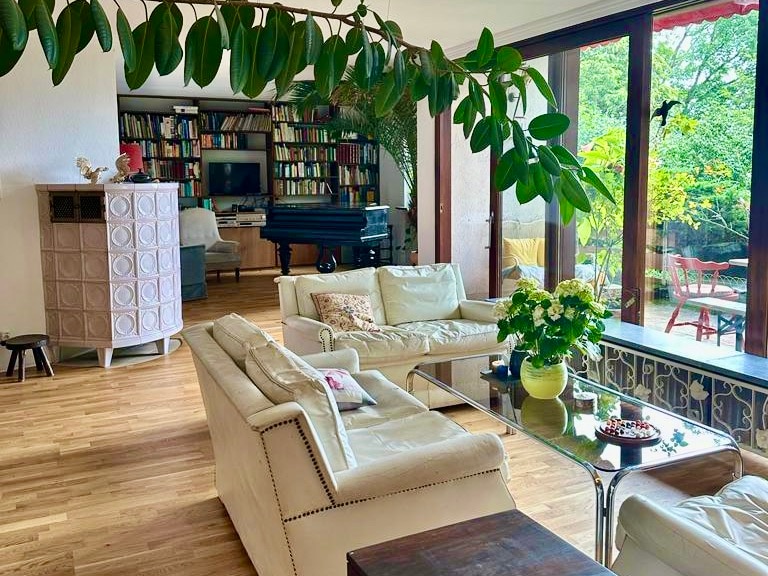 A comfortable living area features soft cream sofas arranged around a glass coffee table. A flowering plant adds a refreshing touch. Panoramic windows provide a view of the garden, while bookshelves can be seen in the background, contributing to a cozy atmosphere.