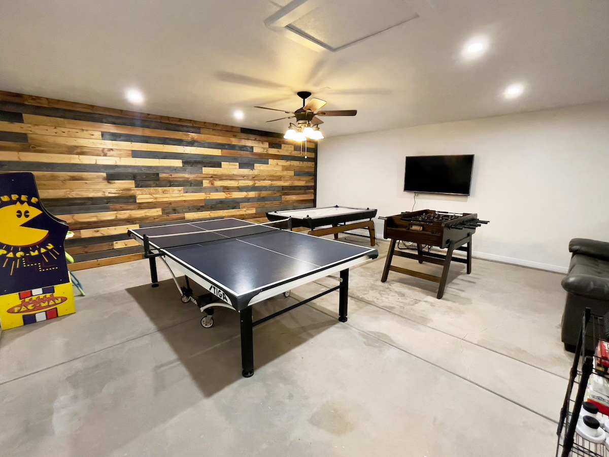 The game room features an air hockey table and a foosball table, complemented by a ping pong table in the center. A wooden wall adds character, while a television is mounted above a low table. Lighting is provided by a ceiling fan.