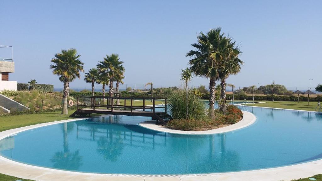 A serene community pool is surrounded by well-maintained greenery and palm trees. A wooden bridge crosses over the water, adding an inviting element to the landscape. The clear blue sky reflects off the calm surface of the pool, enhancing the tranquil setting.