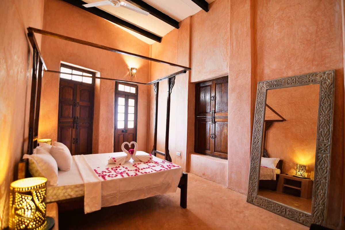 A spacious bedroom features a four-poster bed with a decorated headboard. Soft lighting is provided by lamps on either side, and large wooden windows allow natural light to illuminate the warm, earthy walls. A large mirror and decorative accents enhance the inviting atmosphere.