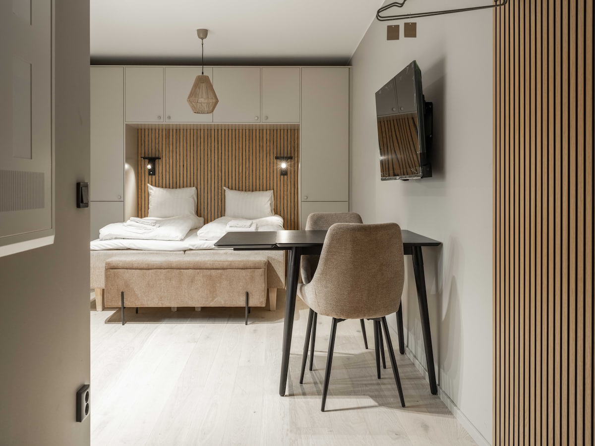A cozy studio features a plush double bed framed by wooden paneling. A compact dining area with a table and two chairs is positioned nearby, along with a wall-mounted television. Soft lighting enhances the modern design throughout the space.