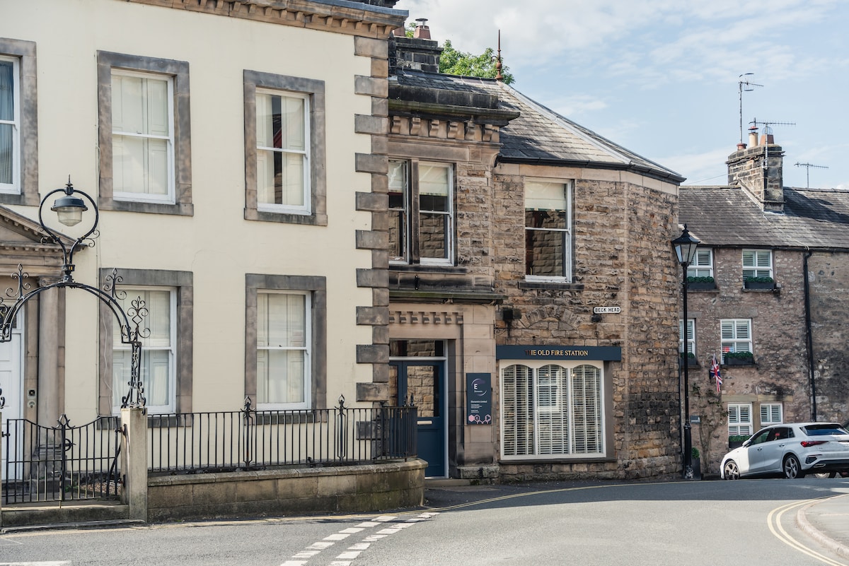 The charming Old Fire Station building features a stone façade and large windows, situated on a quiet corner of the street. The entrance is marked by a sign, while neighboring historic structures hint at the town's heritage.