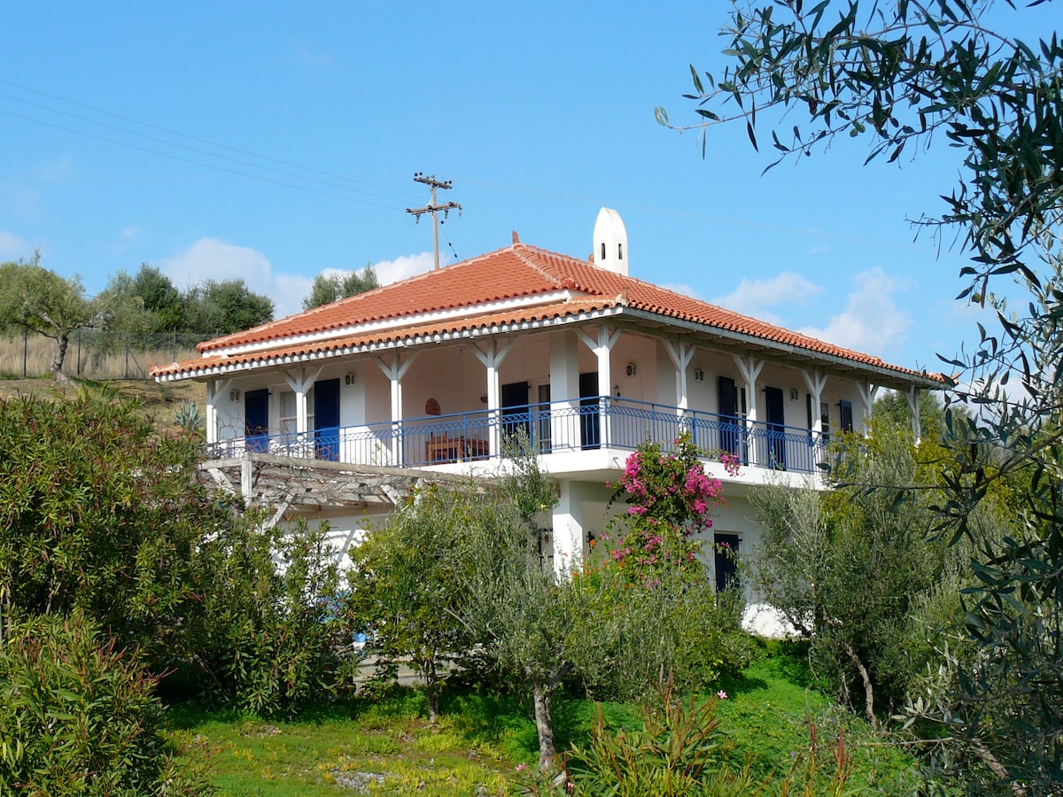 The two-story house showcases traditional Greek architecture with a red-tiled roof and blue shutters. Surrounded by olive trees, it features a spacious balcony on the upper level, offering views of the landscape. Lush greenery and colorful flowers enhance the serene setting.