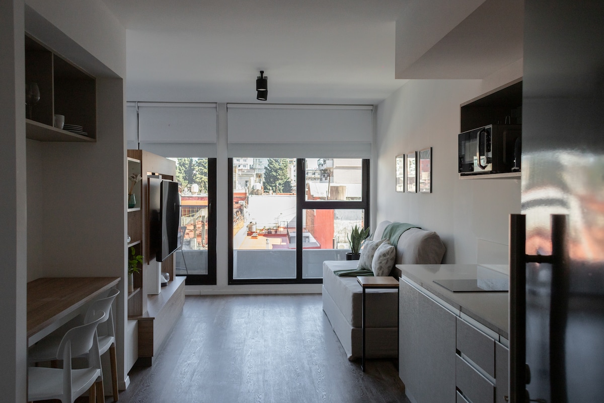 A modern living space features large windows allowing natural light to fill the room. A comfortable seating area is complemented by a dining table and chairs. Minimalist furniture is arranged thoughtfully against the walls, creating an open atmosphere with a stylish design.