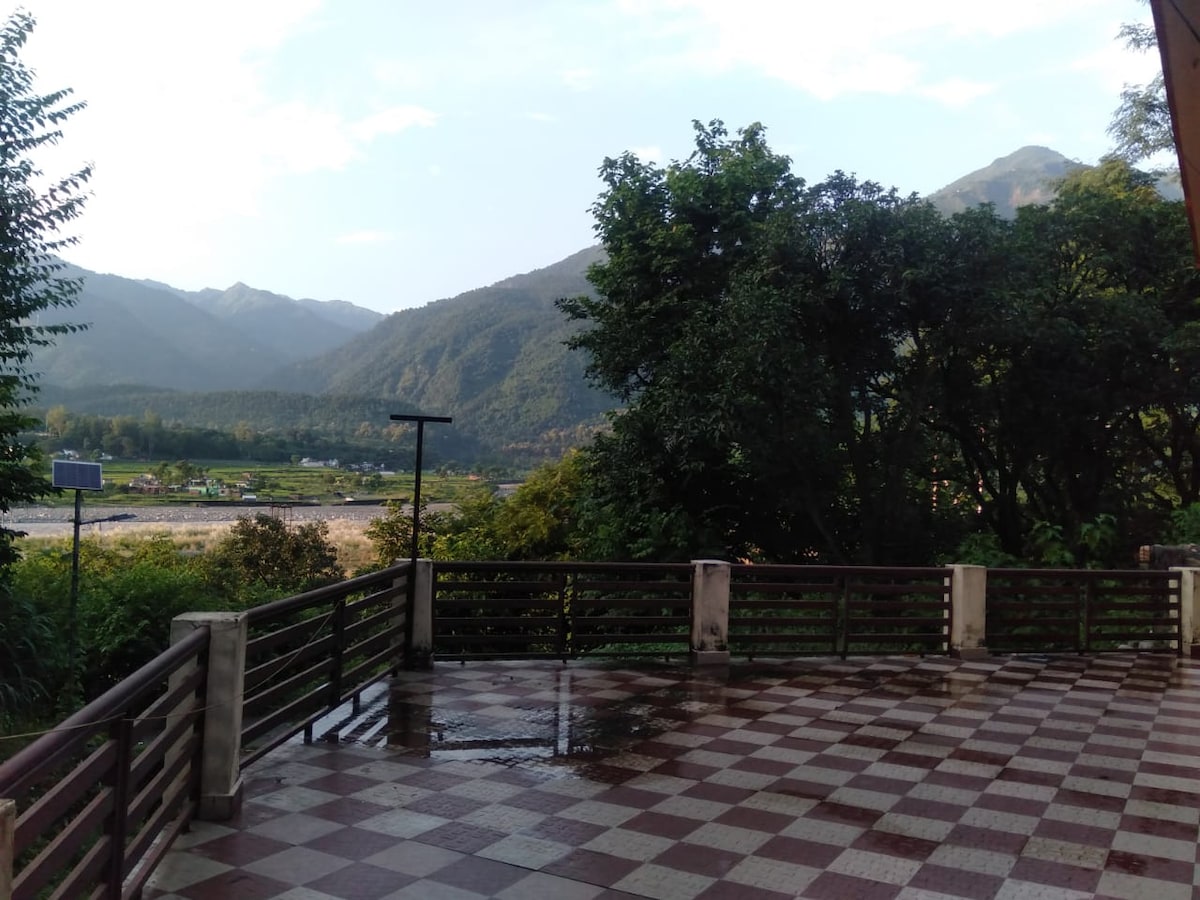 A spacious outdoor balcony is shown, featuring tiled flooring in a checkerboard pattern. Lush greenery and trees are visible, with a backdrop of mountains and the river. The scene captures a tranquil natural environment, ideal for relaxation.