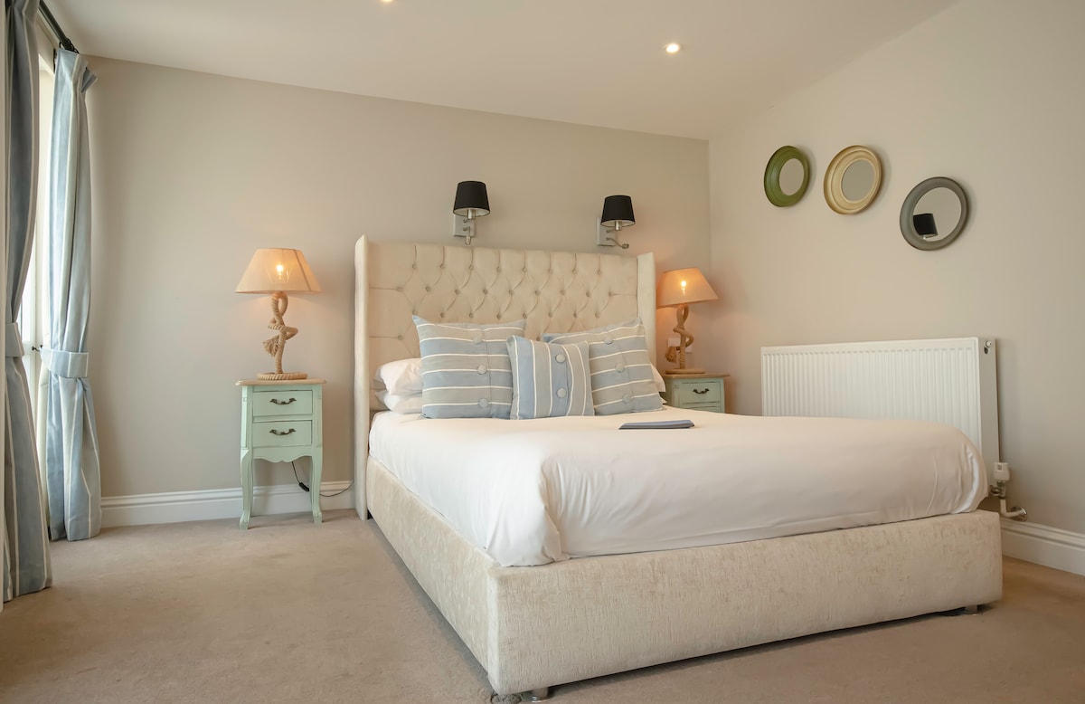 A comfortable bedroom features a spacious bed adorned with soft pillows. Two bedside lamps provide gentle illumination, while stylish wall mirrors and framed circles add a touch of decor. Light curtains frame the window, allowing natural light to fill the room.