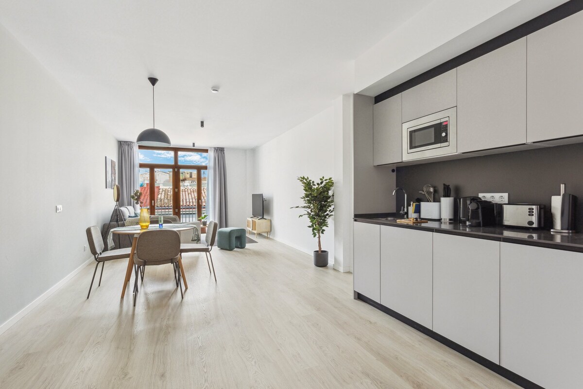 A modern living area is highlighted by a cozy dining table surrounded by four chairs. A sleek kitchen is equipped with essential appliances, while a comfortable seating area includes a TV and windows that lead to a balcony, allowing natural light to fill the space.