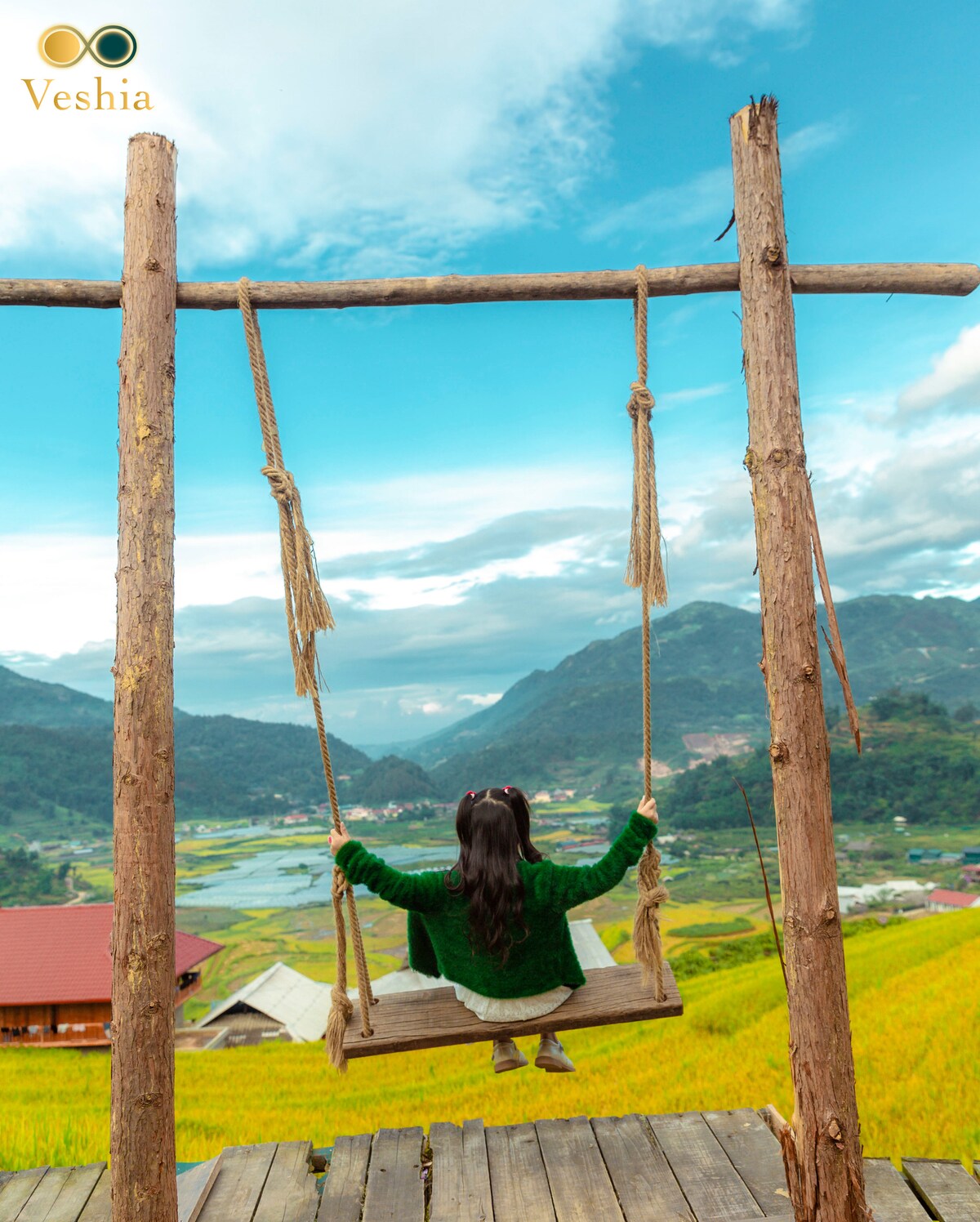 A wooden swing is suspended from sturdy posts, offering a view of lush green mountains and expansive fields below. The sky is bright with scattered clouds, enhancing the natural beauty of the landscape. A person seated on the swing faces the scenic vista.
