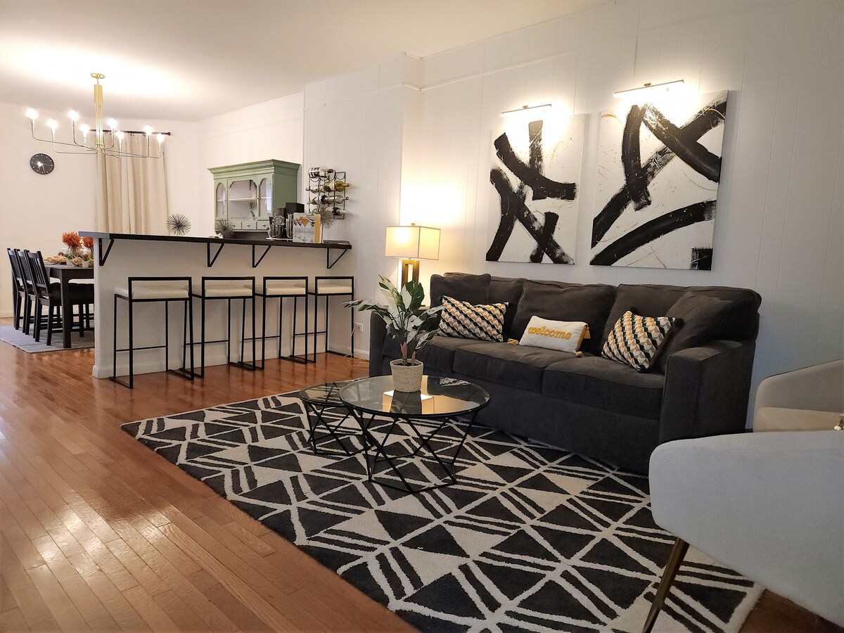 The living area offers a cozy sofa set against a bright, open space. A geometric area rug defines the seating area, complemented by a glass coffee table. Light wood floors create an inviting atmosphere, while a dining area in the background is visible, featuring a dark wooden table.