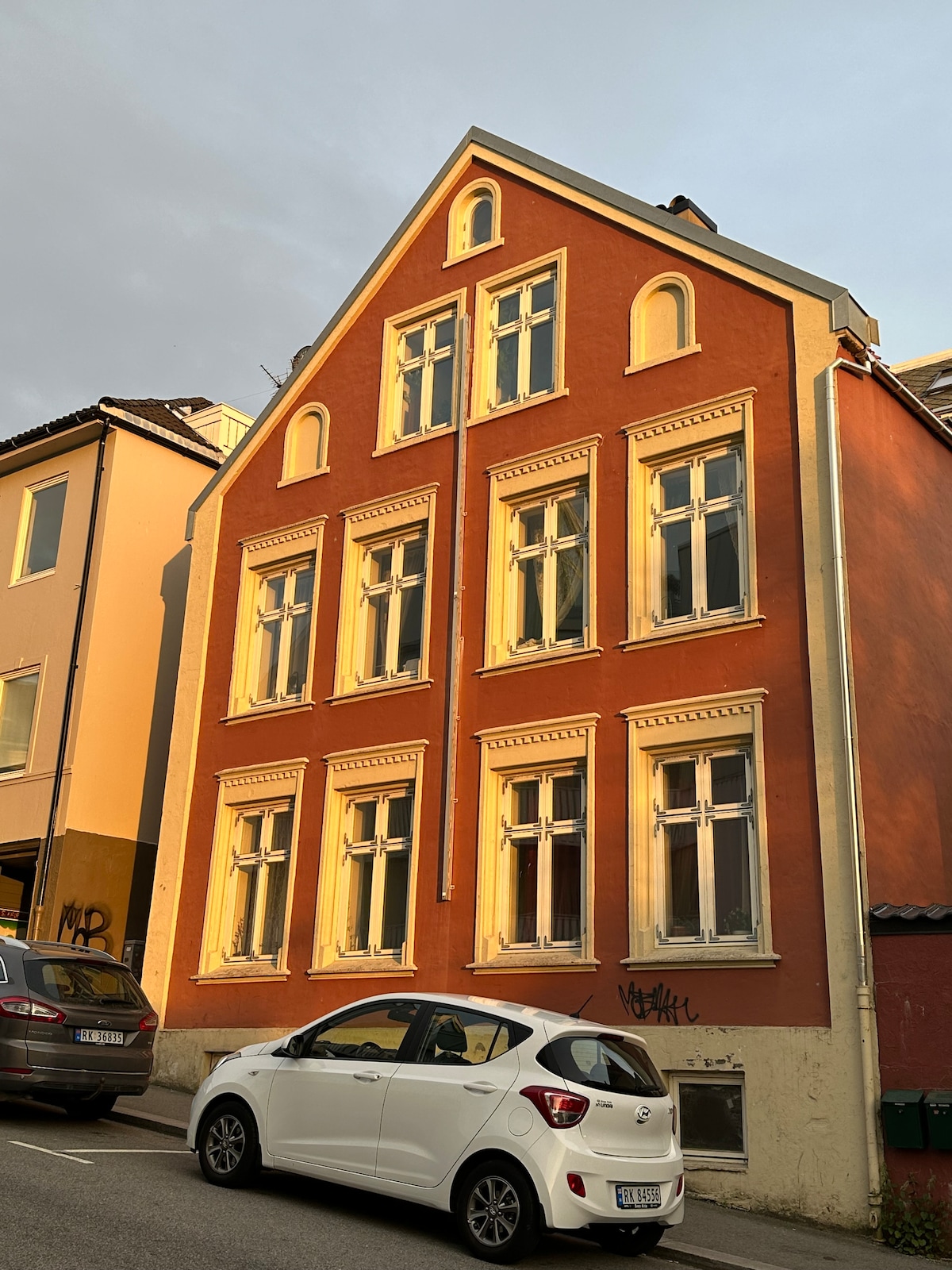A charming historical building with a striking red exterior, featuring large, tall windows that catch the evening light. The structure showcases a unique architectural style with multiple levels and decorative details, creating an inviting presence in the heart of town.
